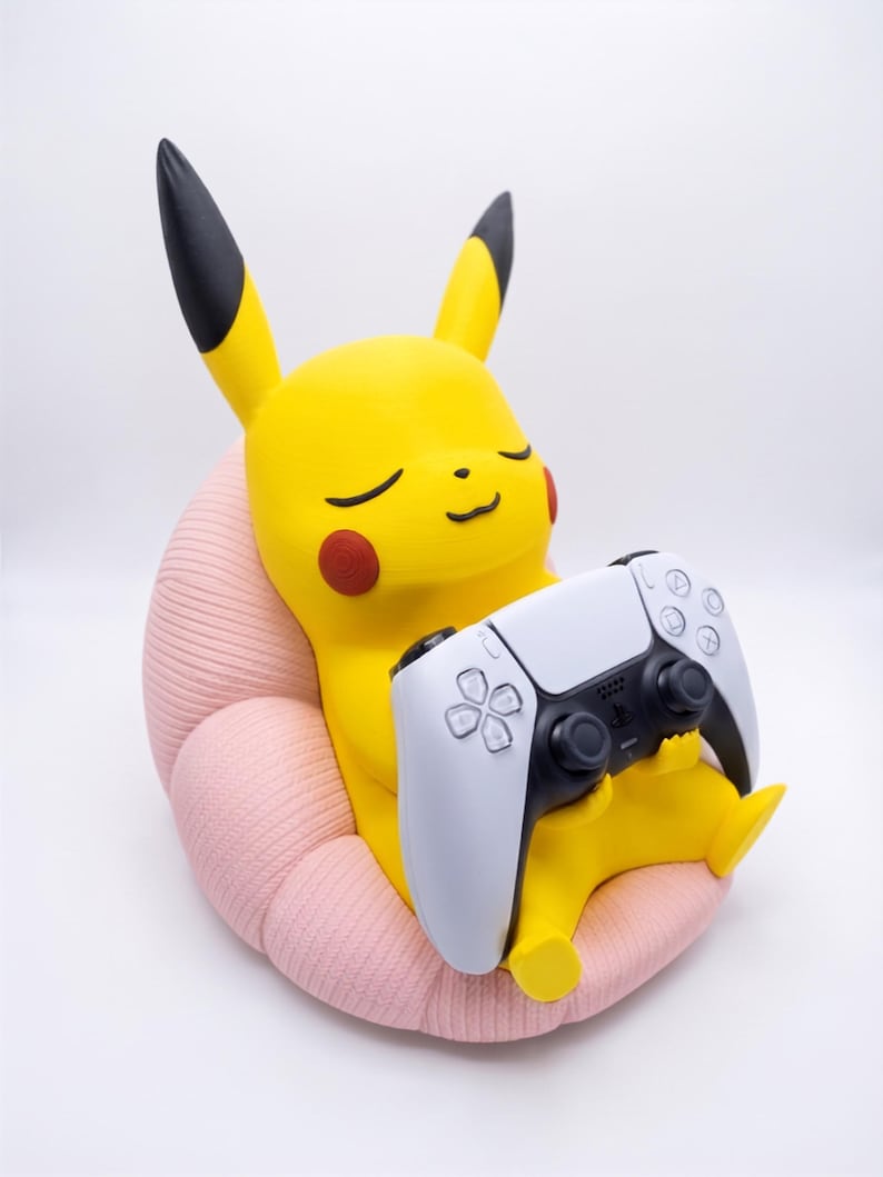May include: A yellow Pikachu figure with black-tipped ears and red cheeks, sitting in a textured light pink chair. The figure holds a white and black wireless game controller in its hands, with its eyes closed in a relaxed, smiling expression.