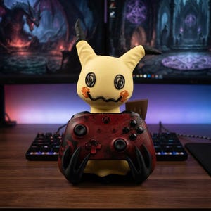 Pode incluir: Um suporte de comando em forma de Mimikyu amarelo com orelhas pretas, olhos em espiral e boca serrilhada, segurando um comando Xbox vermelho e preto com padrões, joysticks e botões pretos nas suas duas mãos em forma de garra.