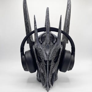 K&ouml;nnte beinhalten: Ein dunkelgrauer Helm mit verwittertem Finish, hohen, spitzen Zacken und komplexen, erhabenen Ornamentmustern. Der Helm wird mit einem Paar schlichter, schwarzer Over-Ear-Kopfh&ouml;rer pr&auml;sentiert, die seitlich &uuml;ber den Ohrmuscheln aufliegen.