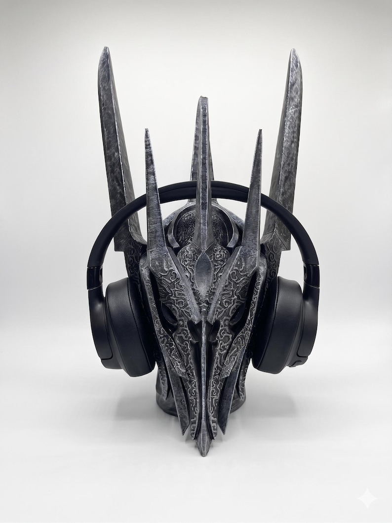 K&ouml;nnte beinhalten: Ein dunkelgrauer Helm mit verwittertem Finish, hohen, spitzen Zacken und komplexen, erhabenen Ornamentmustern. Der Helm wird mit einem Paar schlichter, schwarzer Over-Ear-Kopfh&ouml;rer pr&auml;sentiert, die seitlich &uuml;ber den Ohrmuscheln aufliegen.