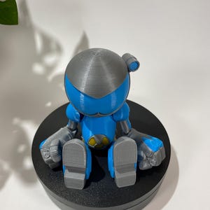 Puede incluir: Una figura de robot impresa en 3D azul y gris sentada sobre una base circular negra. El robot tiene una cabeza redonda con la parte superior gris y una visera azul. El robot est&aacute; sentado con las piernas cruzadas y los brazos apoyados en las rodillas.