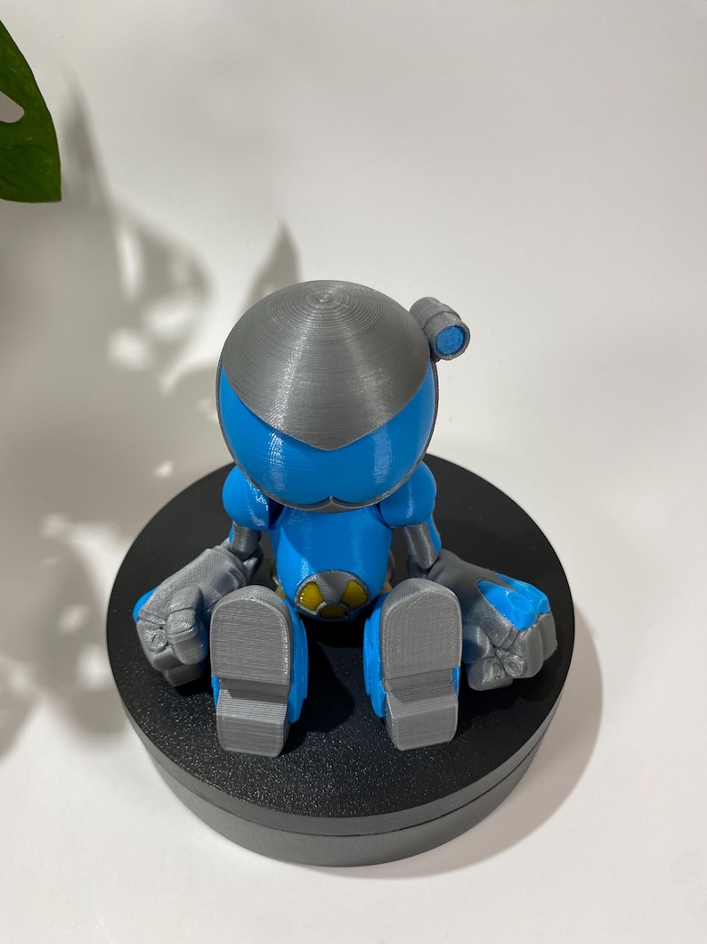 Puede incluir: Una figura de robot impresa en 3D azul y gris sentada sobre una base circular negra. El robot tiene una cabeza redonda con la parte superior gris y una visera azul. El robot est&aacute; sentado con las piernas cruzadas y los brazos apoyados en las rodillas.