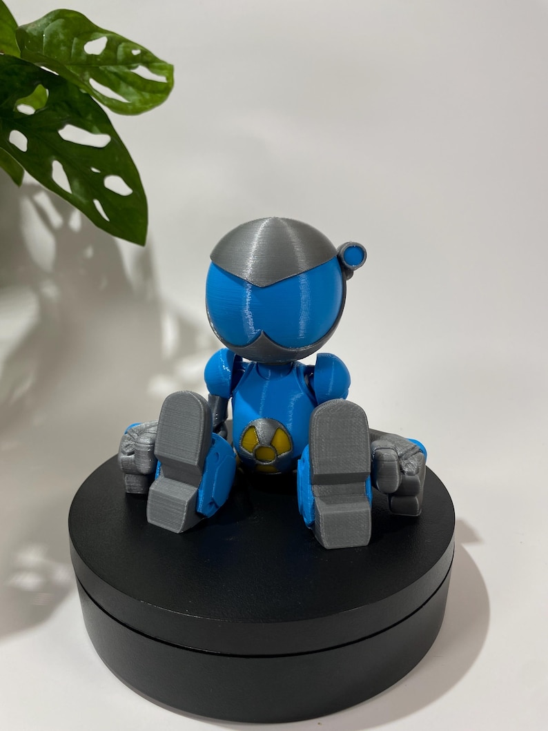 Puede incluir: Una figura de robot impresa en 3D de color azul y plateado sentada sobre una base circular negra. El robot tiene un s&iacute;mbolo amarillo en el pecho.