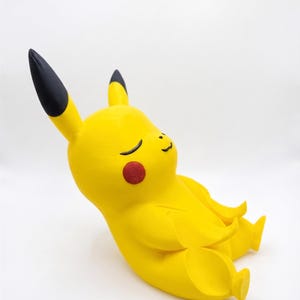 May include: A yellow Pikachu figurine in a seated, meditative pose with eyes closed and a small smile. The character features long ears with black tips, round red cheeks, and a smooth, matte finish, captured from a side-angle perspective.