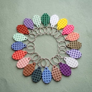 May include: A collection of colorful keychains arranged in a circle. Each keychain features a textured, oval-shaped charm in various colors, including red, purple, green, and yellow, attached to a silver metal ring.