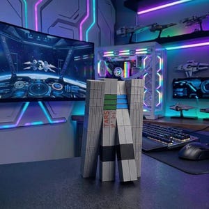 May include: A gray, geometric TARS robot model with green and blue accents, and the word "TARS" printed on the side. The model is on a desk with a computer monitor displaying a spaceship scene, and a computer tower in the background.