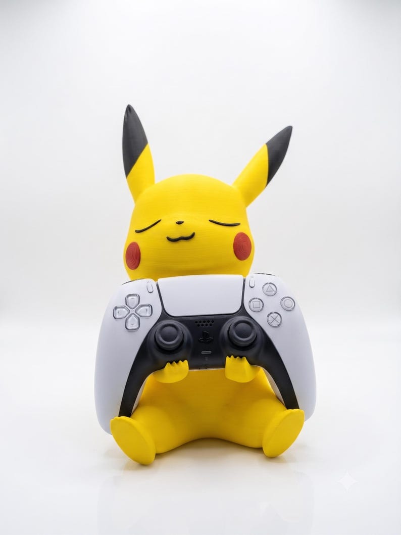 May include: A yellow Pikachu figure with black-tipped ears and red cheeks, sitting in a seated position while holding a white and black video game controller. The figure has a smooth, matte finish and a serene expression with closed eyes.