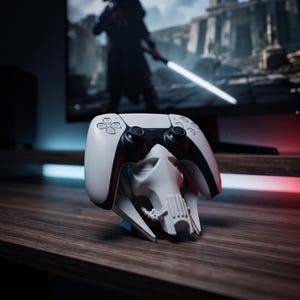 Könnte beinhalten: Ein weiß-schwarzer kabelloser Videospiel-Controller, der auf einem weißen, skulptierten Ständer in Form einer Maske mit mehreren Augen ruht. Der Controller verfügt über zwei Analogsticks, ein Steuerkreuz und Aktionstasten, während der Ständer eine detaillierte, kantige, roboterartige Gesichtsstruktur mit einer markanten zentralen Nase aufweist.