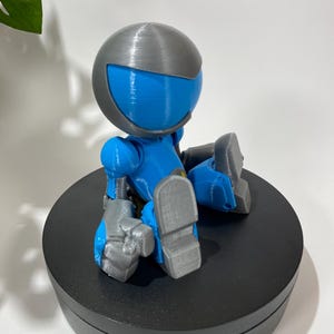 Puede incluir: Una peque&ntilde;a figura de robot azul y gris sentada sobre una base circular negra. El robot tiene una cabeza redonda con un casco gris y una visera azul. El robot est&aacute; sentado con las piernas cruzadas y los brazos cruzados.