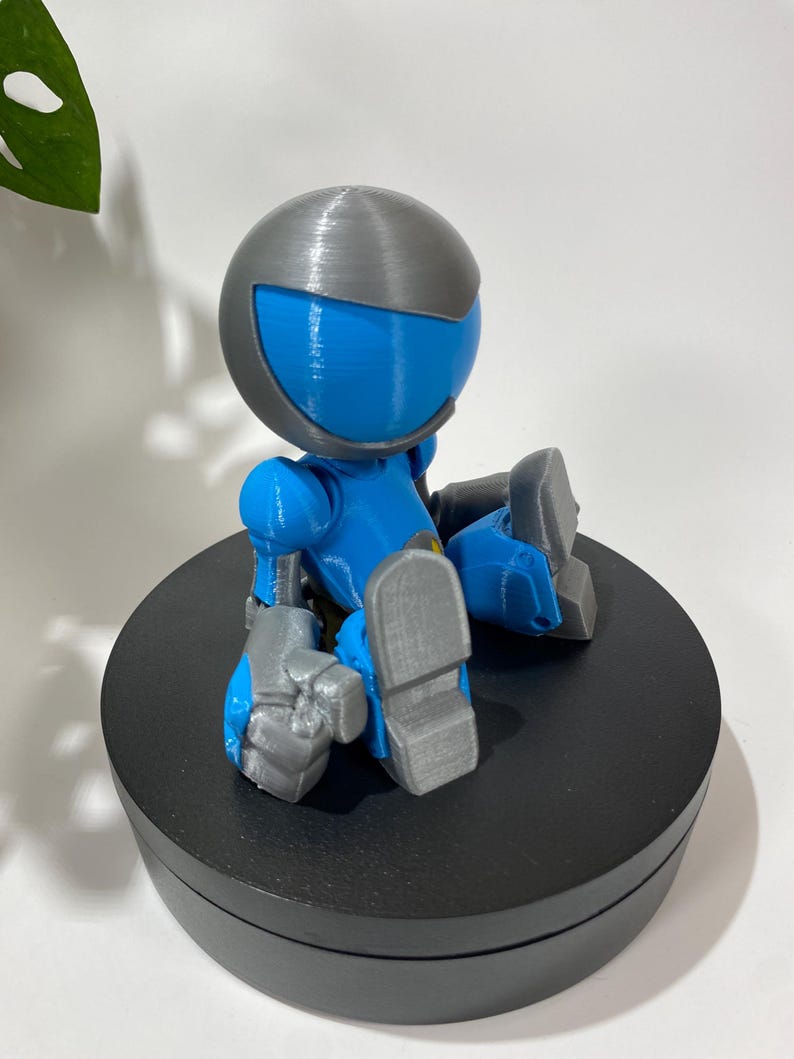 Puede incluir: Una peque&ntilde;a figura de robot azul y gris sentada sobre una base circular negra. El robot tiene una cabeza redonda con un casco gris y una visera azul. El robot est&aacute; sentado con las piernas cruzadas y los brazos cruzados.