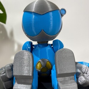 Puede incluir: Una figura de robot impresa en 3D de color azul y gris sentada con las piernas cruzadas. El robot tiene una cabeza grande con una cara azul y un casco gris. El robot lleva un s&iacute;mbolo amarillo y negro en el pecho.