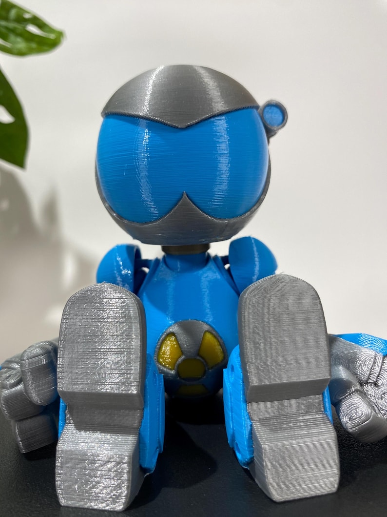Puede incluir: Una figura de robot impresa en 3D de color azul y gris sentada con las piernas cruzadas. El robot tiene una cabeza grande con una cara azul y un casco gris. El robot lleva un s&iacute;mbolo amarillo y negro en el pecho.