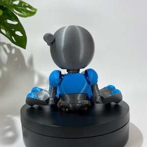 Puede incluir: Una peque&ntilde;a figura de robot gris y azul sentada sobre una base circular negra. El robot tiene una cabeza redonda y est&aacute; sentado con las piernas cruzadas.