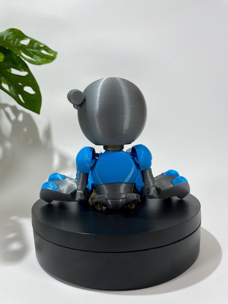 Puede incluir: Una peque&ntilde;a figura de robot gris y azul sentada sobre una base circular negra. El robot tiene una cabeza redonda y est&aacute; sentado con las piernas cruzadas.