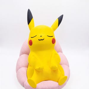 May include: A yellow Pikachu figure with black-tipped ears and red cheeks, depicted with closed eyes and a calm expression, sitting upright in a soft, light pink, ribbed, rounded chair-like cushion against a plain white background.