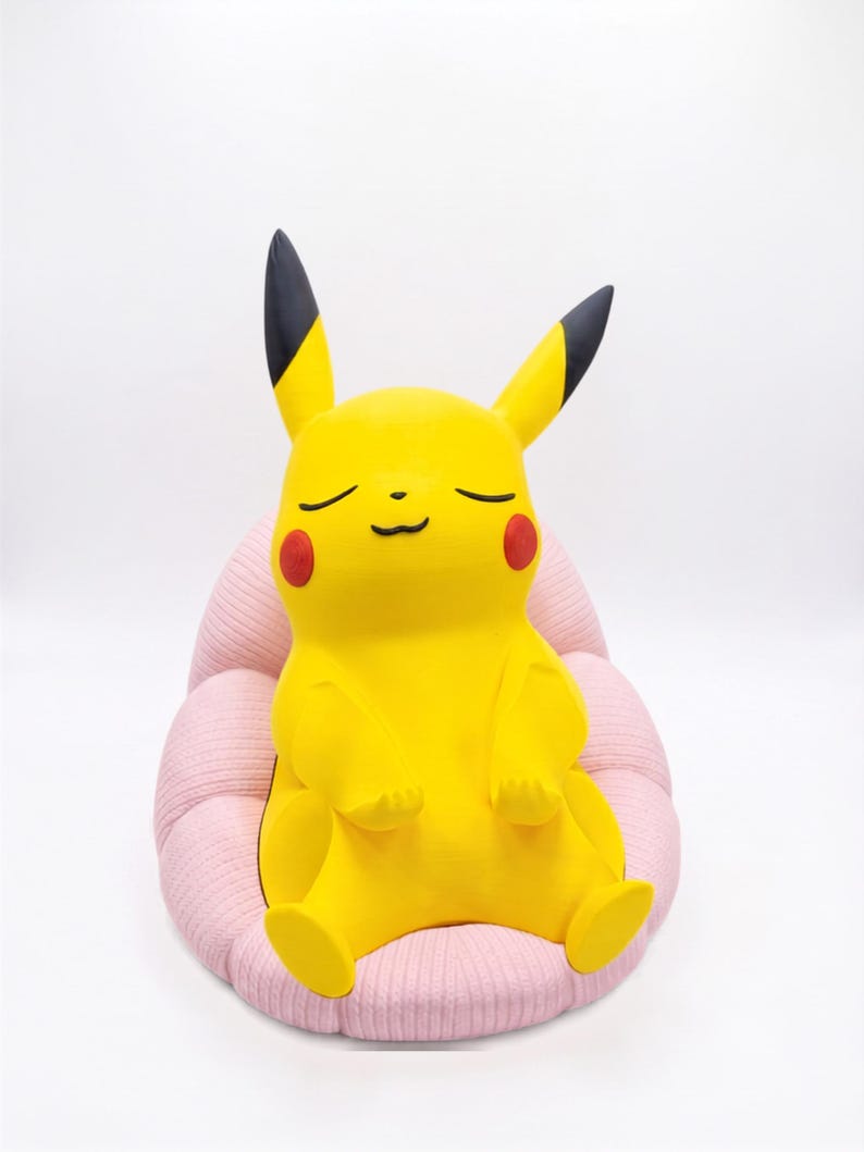 May include: A yellow Pikachu figure with black-tipped ears and red cheeks, depicted with closed eyes and a calm expression, sitting upright in a soft, light pink, ribbed, rounded chair-like cushion against a plain white background.