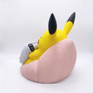 May include: A yellow Pikachu figure with black-tipped ears sitting in a light pink, textured beanbag chair. The figure is holding a white and black game controller in its hands, positioned as if playing a video game while seated in the chair.