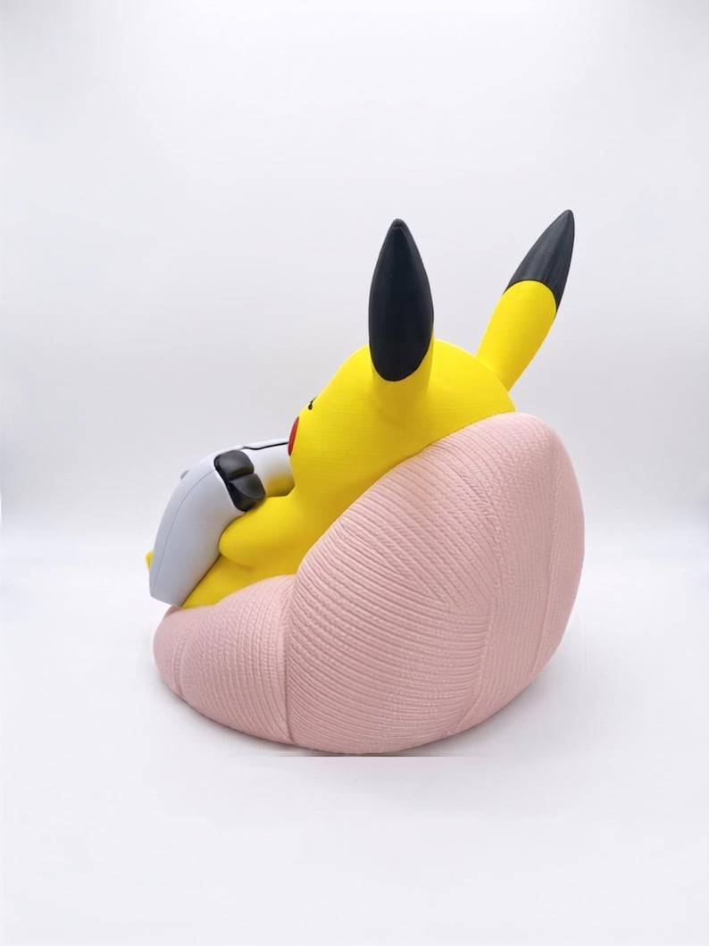 May include: A yellow Pikachu figure with black-tipped ears sitting in a light pink, textured beanbag chair. The figure is holding a white and black game controller in its hands, positioned as if playing a video game while seated in the chair.
