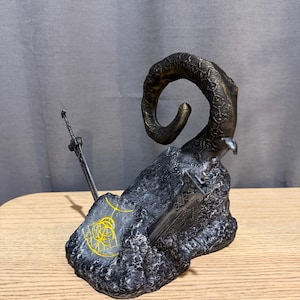 Op de afbeelding: Een gedetailleerde sculptuur met een grote, gebogen hoorn met een textuur, die oprijst uit een donkere, rotsachtige basis. Twee zwaarden zijn in de basis ingebed. Een gouden ontwerp is zichtbaar op de basis. De sculptuur staat op een houten oppervlak.