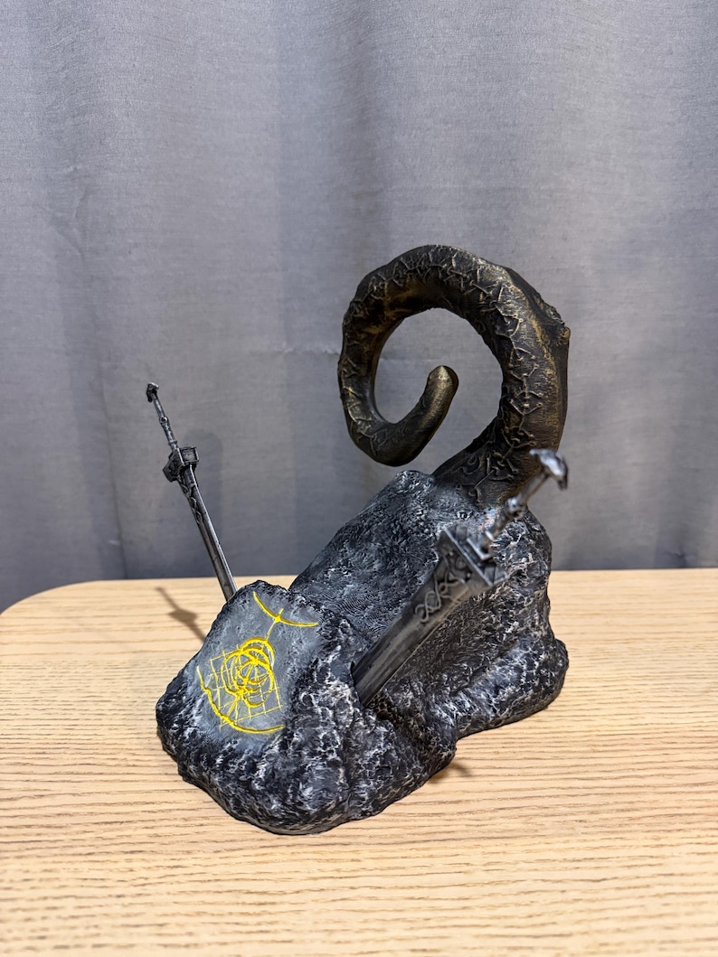 Op de afbeelding: Een gedetailleerde sculptuur met een grote, gebogen hoorn met een textuur, die oprijst uit een donkere, rotsachtige basis. Twee zwaarden zijn in de basis ingebed. Een gouden ontwerp is zichtbaar op de basis. De sculptuur staat op een houten oppervlak.