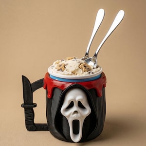 May include: A black mug with a white Ghostface mask design, a red drip effect, and a knife-shaped handle. The mug is filled with ice cream and two silver spoons.