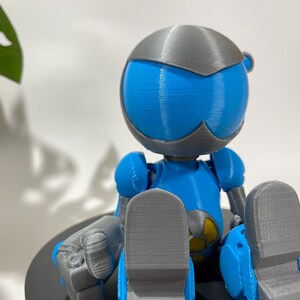 Puede incluir: Una figura de robot impresa en 3D de color azul y gris sentada con las piernas cruzadas. El robot tiene una cabeza redonda con una visera gris y un s&iacute;mbolo amarillo en el pecho.