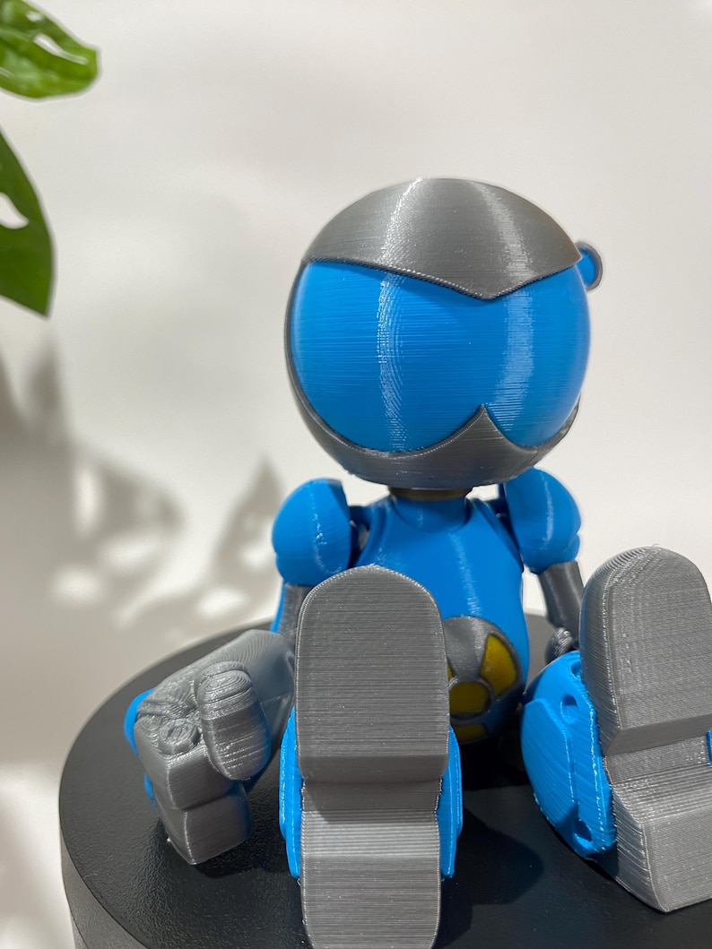 Puede incluir: Una figura de robot impresa en 3D de color azul y gris sentada con las piernas cruzadas. El robot tiene una cabeza redonda con una visera gris y un s&iacute;mbolo amarillo en el pecho.