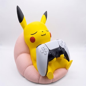 May include: A yellow Pikachu figure with black-tipped ears and red cheeks sitting in a textured light pink beanbag chair. The figure is holding a white and black video game controller with two analog sticks and various buttons in its hands.
