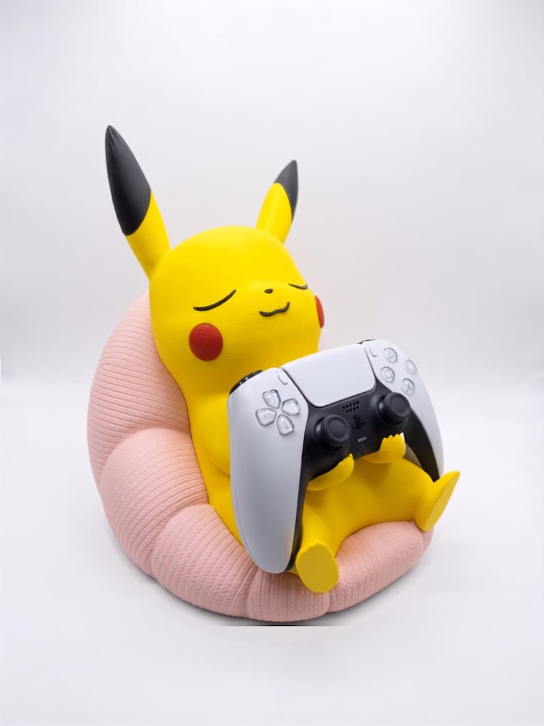 May include: A yellow Pikachu figure with black-tipped ears and red cheeks sitting in a textured light pink beanbag chair. The figure is holding a white and black video game controller with two analog sticks and various buttons in its hands.
