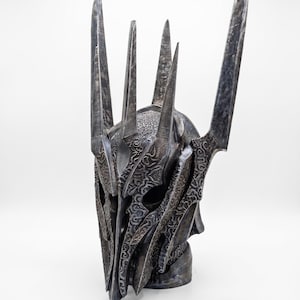 K&ouml;nnte beinhalten: Ein dunkler Metallhelm mit einer hohen, spitzen Krone und mehreren vertikalen Zacken. Die Oberfl&auml;che ist mit komplizierten, erhabenen Schn&ouml;rkelmustern bedeckt und hat ein verwittertes, gunmetallgraues Finish mit tiefen Schatten.
