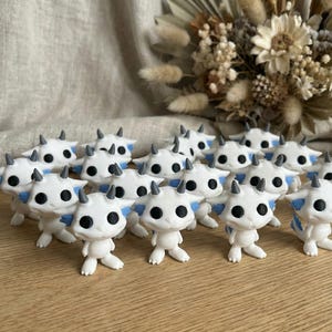 Puede incluir: Una colección de pequeñas figuras blancas con ojos negros, cuernos grises y detalles azules. Las figuras están dispuestas sobre una superficie de madera, con un arreglo floral seco en el fondo. Las figuras parecen criaturas de fantasía.