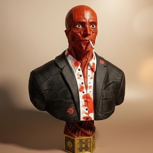 Puede incluir: Escultura de busto de piel roja de un hombre con blazer negro, camisa blanca con salpicaduras rojas y cigarrillo en la boca. El busto se asienta sobre una base decorativa dorada y marrón. La escultura tiene una apariencia muscular detallada.