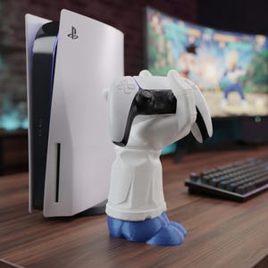 Puede incluir: Una figura blanca y azul con forma de manos que sostienen un mando de juego blanco y negro, colocada frente a una consola de juegos blanca. La figura presenta una sección superior blanca y una base azul, diseñada para exhibir el mando.