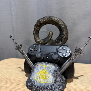 Op de afbeelding: Een zwarte PlayStation 4-controller staat centraal in deze afbeelding, geflankeerd door twee zilveren zwaarden en een steen met een gouden embleem. Een bronskleurige, gebogen hoorn rijst op achter de controller, wat een uniek gaming-accessoire cre&euml;ert.