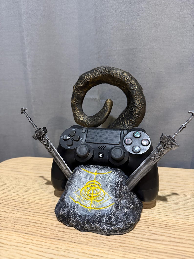Op de afbeelding: Een zwarte PlayStation 4-controller staat centraal in deze afbeelding, geflankeerd door twee zilveren zwaarden en een steen met een gouden embleem. Een bronskleurige, gebogen hoorn rijst op achter de controller, wat een uniek gaming-accessoire cre&euml;ert.