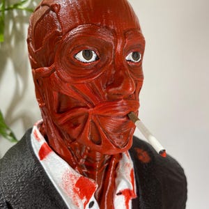 Pu&ograve; includere: Una scultura anatomica rossa di una testa umana, con muscoli e vene visibili. La figura indossa una giacca nera e una camicia bianca con schizzi rossi. Una sigaretta accesa &egrave; in bocca. La scultura ha un aspetto dettagliato e strutturato.