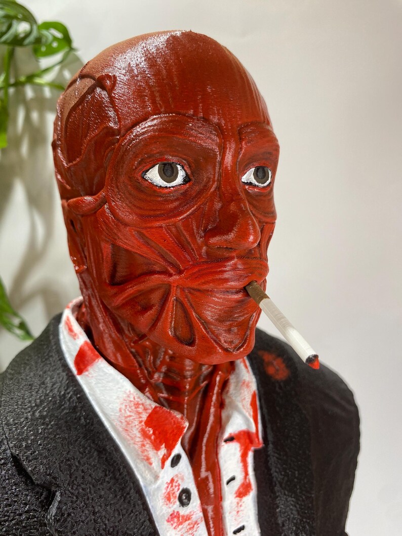 Pu&ograve; includere: Una scultura anatomica rossa di una testa umana, con muscoli e vene visibili. La figura indossa una giacca nera e una camicia bianca con schizzi rossi. Una sigaretta accesa &egrave; in bocca. La scultura ha un aspetto dettagliato e strutturato.