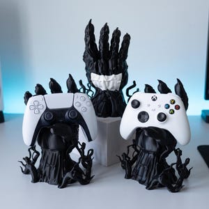 May include: Three black, textured, claw-like stands designed to hold gaming controllers. Two stands hold white game controllers, one with a PlayStation layout and one with an Xbox layout. A third, taller stand sits in the back center.