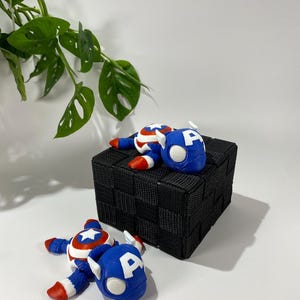 May include: Two Captain America figurines in a playful pose, with blue helmets featuring a white "A" and red, white, and blue costumes. They are placed on a black woven cube against a white backdrop, with green leafy plant in the background.