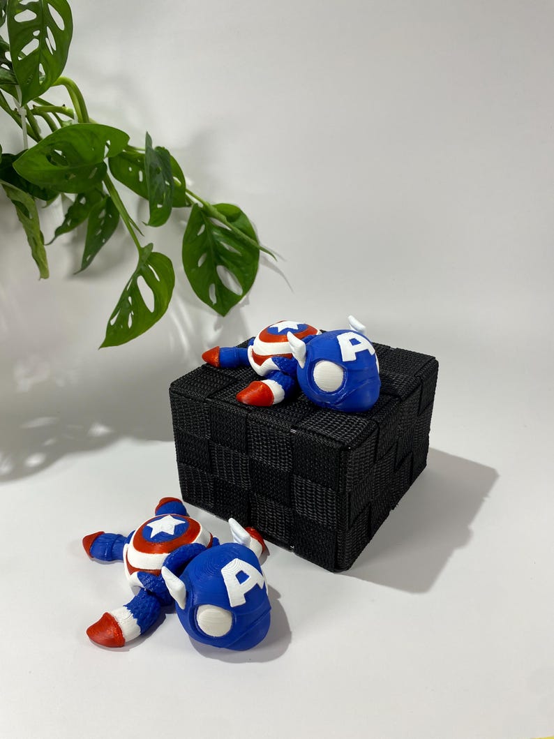 May include: Two Captain America figurines in a playful pose, with blue helmets featuring a white "A" and red, white, and blue costumes. They are placed on a black woven cube against a white backdrop, with green leafy plant in the background.