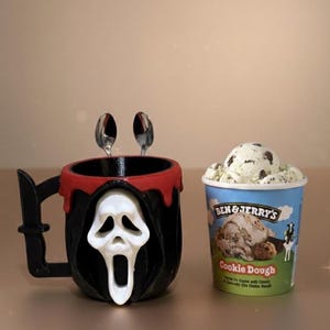 Può includere: Una tazza nera e rossa a tema Halloween con un design a maschera Ghostface bianca e un manico a forma di coltello. Due cucchiaini sono all'interno della tazza. Accanto, una vaschetta di gelato Ben & Jerry's Cookie Dough.