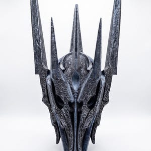 K&ouml;nnte beinhalten: Ein dunkelgrauer, metallisch wirkender Helm mit einer hohen, spitzen Krone und mehreren vertikalen Zacken. Die Oberfl&auml;che ist mit komplexen, erhabenen Wirbelmustern und Texturen bedeckt, die ihm ein gealtertes, verwittertes Aussehen mit scharfen Kanten verleihen.