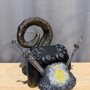 Op de afbeelding: Een zwarte PlayStation 4-controller rust op een grijs en zwarte rotsachtige basis, geflankeerd door twee zilveren zwaarden. Een grote, gebogen, bronskleurige hoorn rijst op achter de controller. De basis heeft een geel embleem.