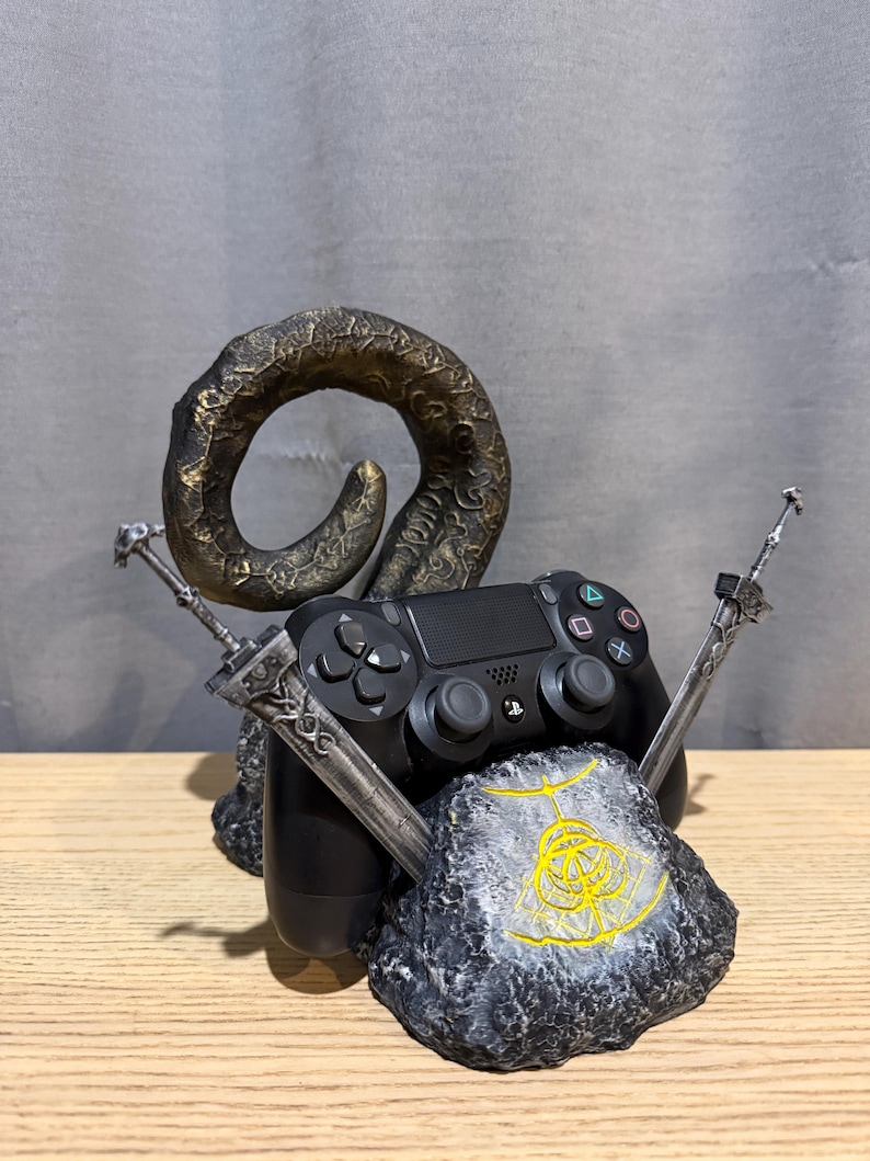 Op de afbeelding: Een zwarte PlayStation 4-controller rust op een grijs en zwarte rotsachtige basis, geflankeerd door twee zilveren zwaarden. Een grote, gebogen, bronskleurige hoorn rijst op achter de controller. De basis heeft een geel embleem.