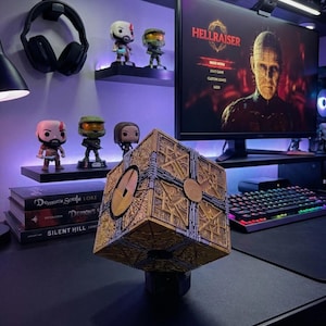 May include: A gold and silver puzzle box, with intricate geometric designs, sits on a desk. The box is the focal point, with a computer monitor displaying "Hellraiser" in the background. Other items include figurines, books, and a keyboard.