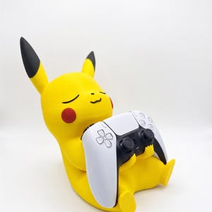 May include: A yellow Pikachu figure with black-tipped ears and red cheeks, depicted in a seated position. The figure is holding a white and black wireless video game controller in its hands, with its eyes closed in a relaxed expression.