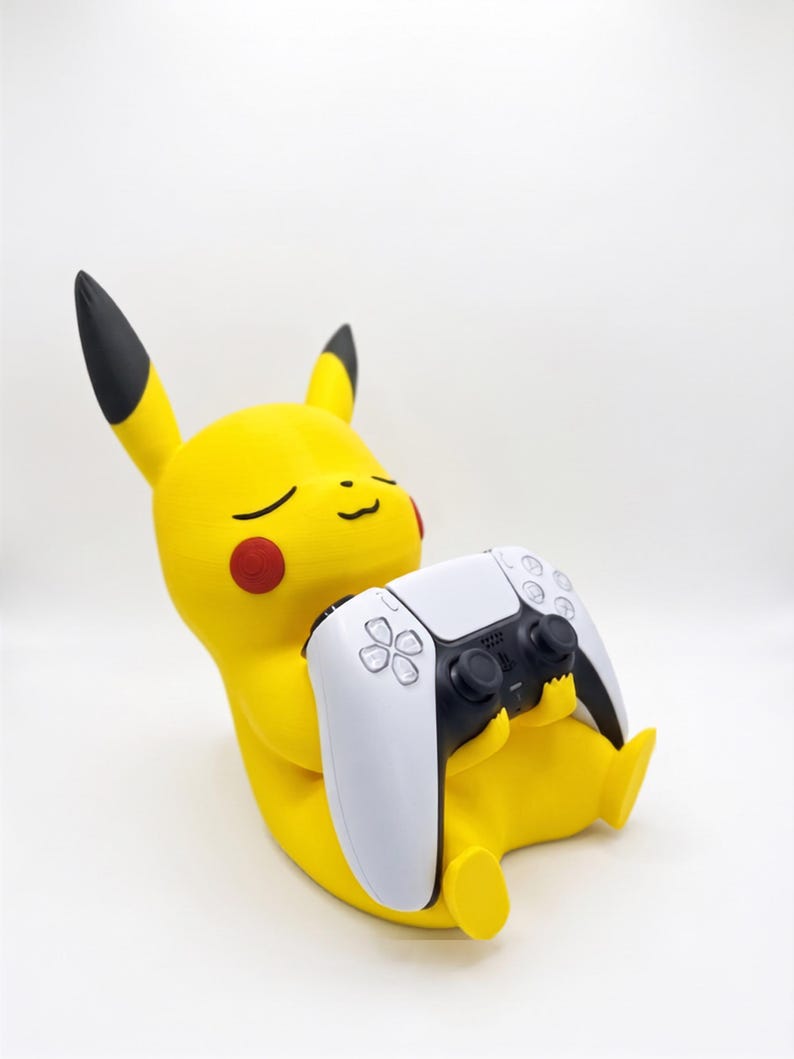 May include: A yellow Pikachu figure with black-tipped ears and red cheeks, depicted in a seated position. The figure is holding a white and black wireless video game controller in its hands, with its eyes closed in a relaxed expression.