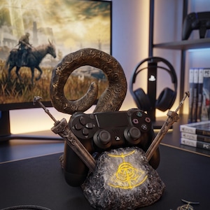 Op de afbeelding: Een zwarte PlayStation 4-controller rust op een decoratieve standaard. De standaard heeft een stenen basis met een gloeiend geel symbool, een gebogen hoorn en twee gekruiste zwaarden. Een gamingmonitor en koptelefoon staan op de achtergrond.