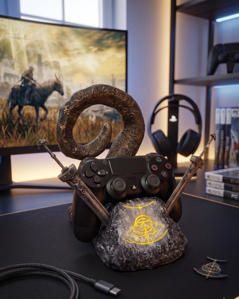 Op de afbeelding: Een zwarte PlayStation 4-controller rust op een decoratieve standaard. De standaard heeft een stenen basis met een gloeiend geel symbool, een gebogen hoorn en twee gekruiste zwaarden. Een gamingmonitor en koptelefoon staan op de achtergrond.