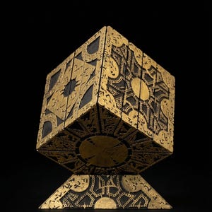 May include: A gold and black puzzle box with intricate geometric patterns. The cube-shaped box sits on a matching gold base, with a dark background. The box has a sunburst design on several faces.